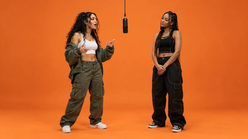 Two women beside a hanging microphone in an orange studio, with the left performer gesturing and the right performer listening